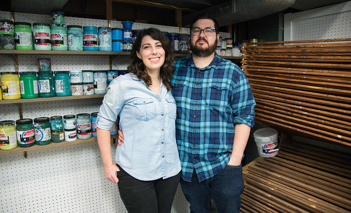 Christina and Dennis Jacobs in their home screen printing studio, standing in front of a rainbow wall of Speedball ink jars