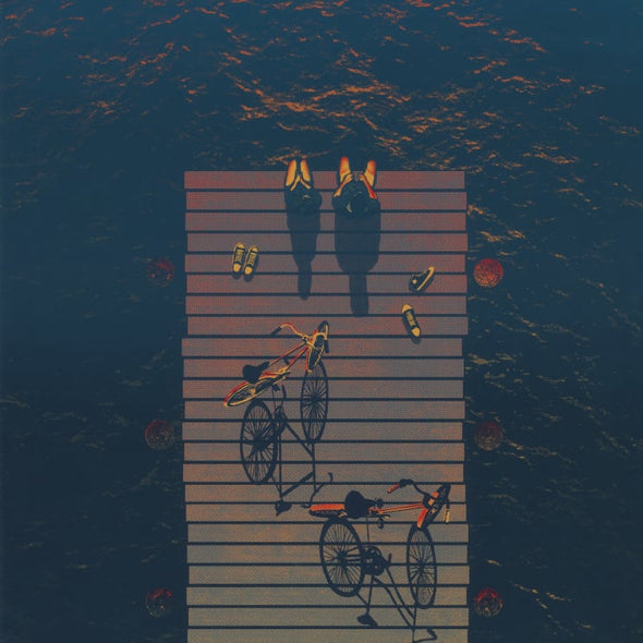 Two people sit on a dock facing the sunset with their bikes behind them.