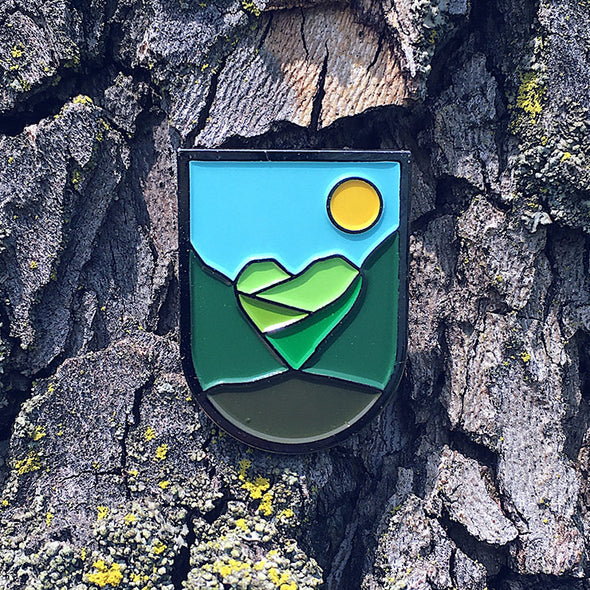 Enamel pin depicting the sky, sun and green hills that form the shape of a heart. 