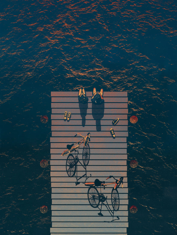 Two people sit on a dock next to their bikes and their shoes as the sun sets across the water.